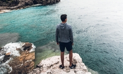 A teen boy standing on a large rock looking out at a body of water.