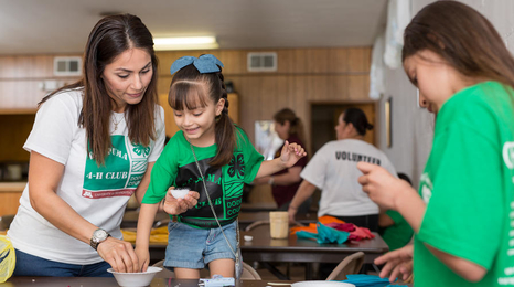 A woman wearing a 4-H shirt helping a young girl with a bow in her hair with an activity. 