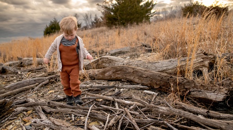 A boy wearing tan overalls walking on a pile of branches in an open pasture.