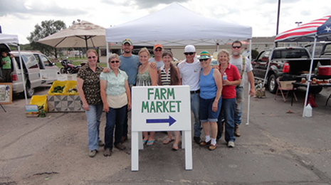Group of people standing together behind a Farm Market sign. Farmers' market booths set up behind the group.