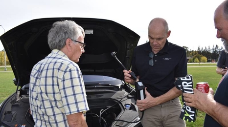 Men observing a demonstration of part of an electric vehicle under the hood.