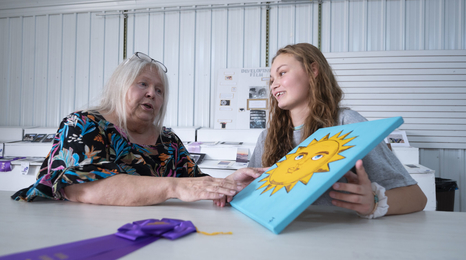 An older woman talking to a young girl about a painting of a sun she is holding.