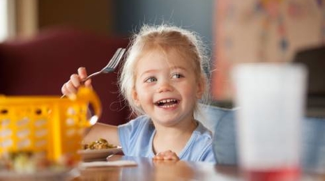 young girl at a table with food on a plate and a fork in her raised hand