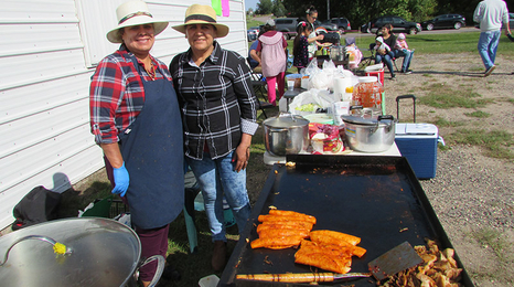 Two female chefs standing behind a large flat top grill with food items being cooked.