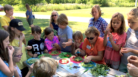 children gathered around a picnic table outside with adults helping prepare vegetables 