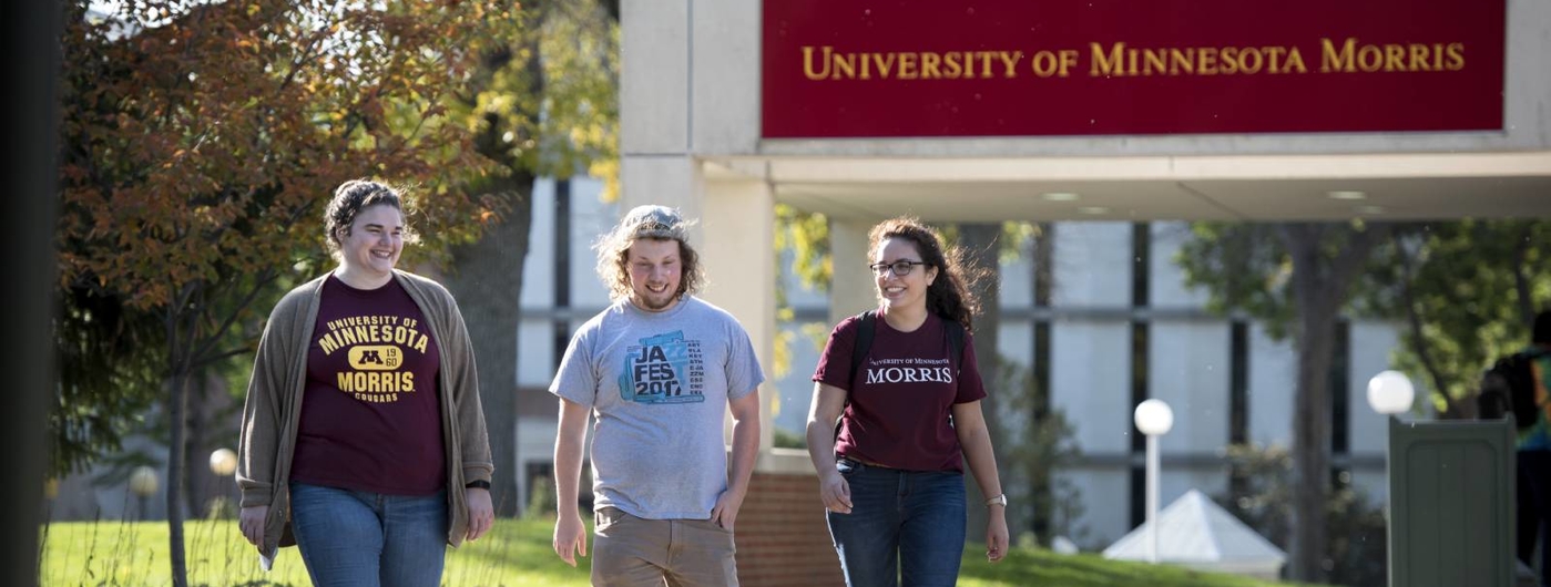 Three students walking on the sidewalk with University of Minnesota Morris sign in the background.