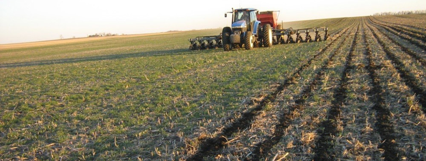 Large blue tractor pulling an implement to cut rows in the soil of a large field.