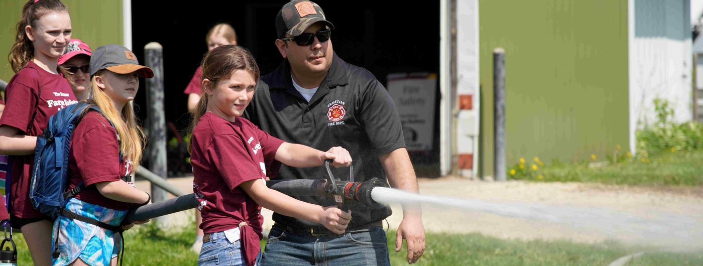 A firefighter with a Preston Fire logo on his shirt teaches a girl around age 12 how to use a fire hose while others wait in line for their turn