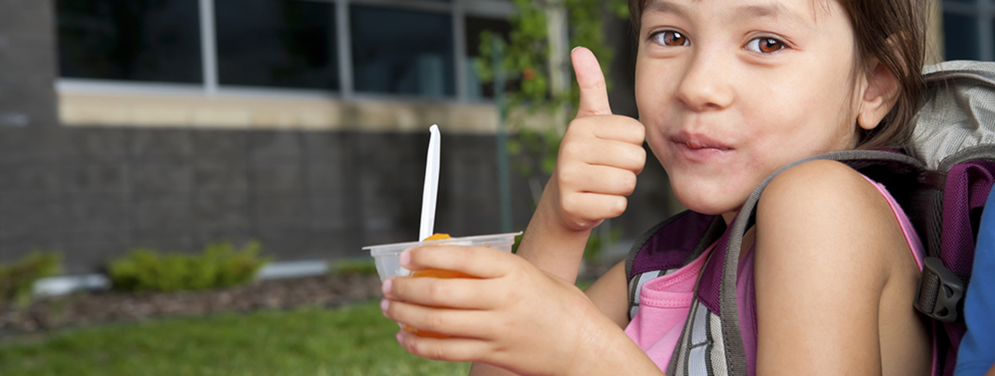 Young girl with backpack eating out of fruit cup and giving thumbs up