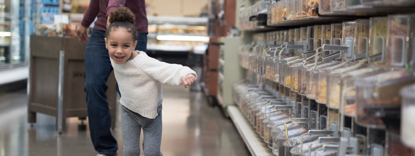 child approximately 8 years old pulls her caregiver or father through the grocery store with a smile on her face. Bulk bins with scoops are on the right.