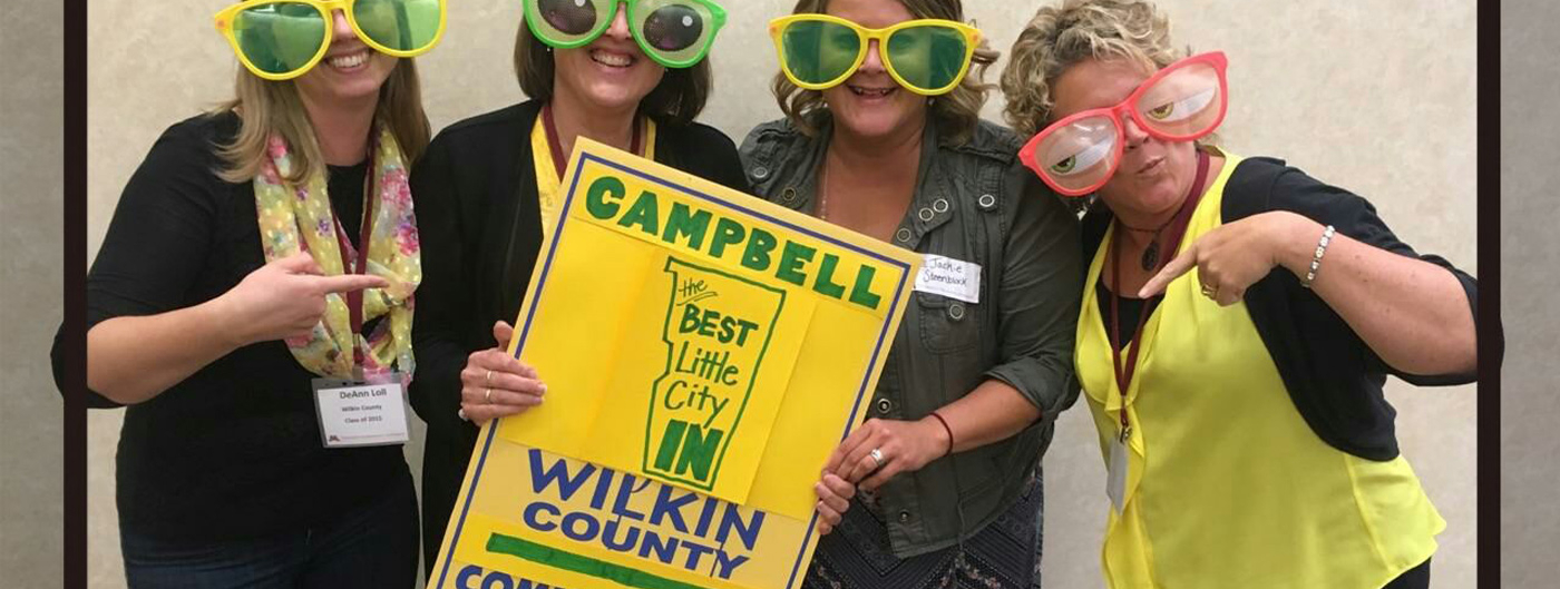 Four ladies wearing fun-oversized glasses holding a sign reading 'Cambell, the best little city in Wilkin County - CommUNITY'