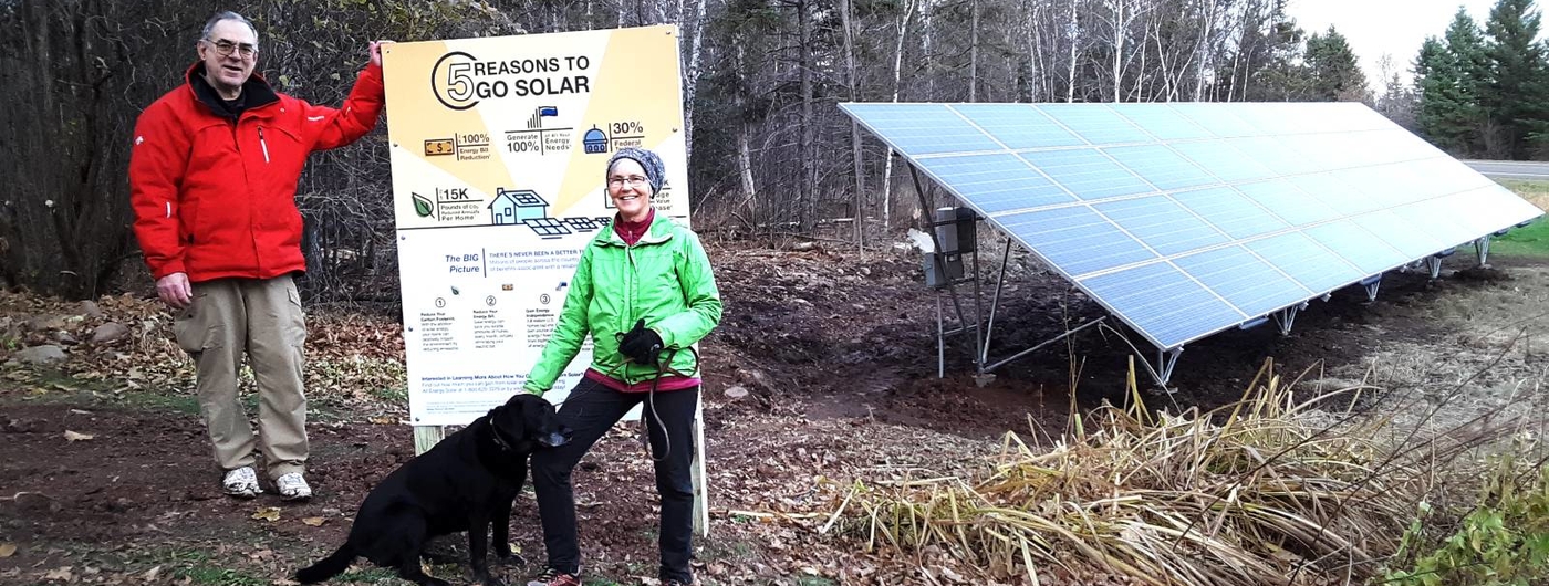 Man, woman and black lab standing next to a solar sign and a photo voltaic solar array.