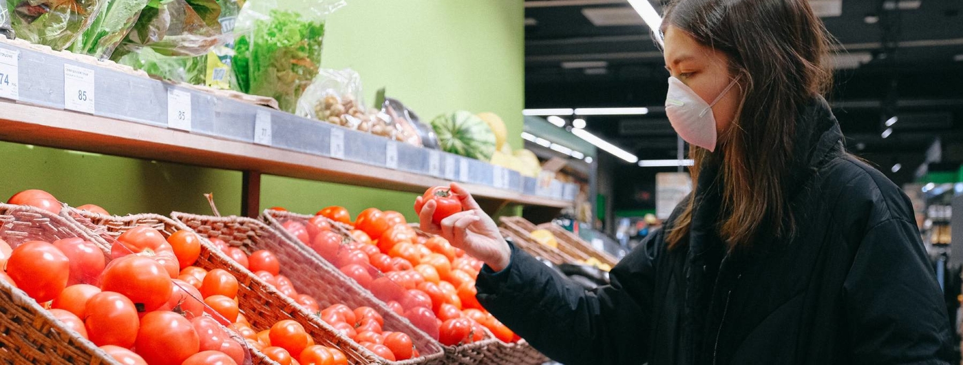 Woman in a grocery store selecting a tomato and wearing a face mask.