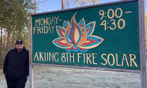Gwe Gasco serves as Sales & Marketing Director for 8th Fire Solar. He stands next to sign that gives Monday through Friday 9 to 4:30 as the hours.