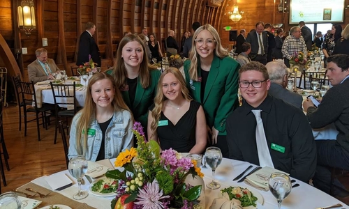 Three youth sitting at a table with official 4-H name tags and two youth standing behind them with green blazers on