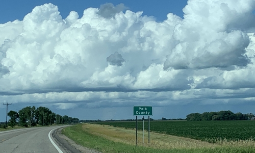 A curved tar road next to trees with a lot of clouds in the sky, and a field with a green road sign in the ditch that says 'Polk County' on it.