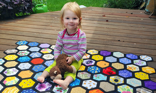 A girl sits on a quilt with a rainbow honeycomb design.