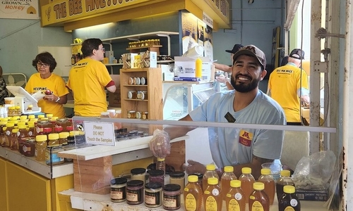 Josh smiles in front of many jars of honey. There is a honey sign behind him and a Minnesota Grown sign on the wall.