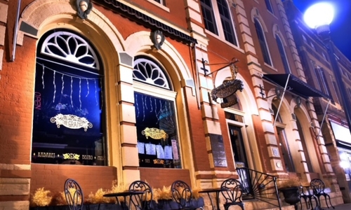 Nighttime front of the historic building with a lantern and patio tables in front. The windows are arched and have decorative metal work at the top.