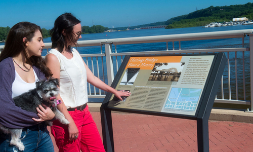Two young women, one holding a small dog, reading an outdoor-historical landmark sign.