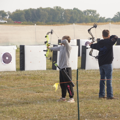 Two boys aiming a bow and arrow at a row of targets.