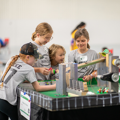 Four girls wearing matching gray team t-shirts work on their Rube Goldberg machine. 