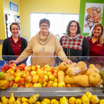 Four educators stand behind a bin of apples and squash in a colorful, well-lit food shelf 