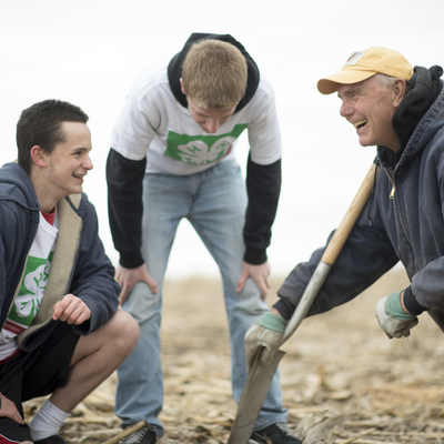 A boy wearing a 4-H t-shirt crouching in a field talking to a man wearing a baseball cap crouching with a shovel while another boy standing looks down 
