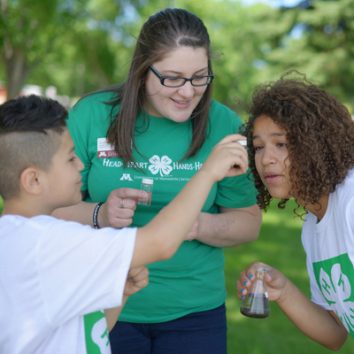 A woman in a green shirt looking at a boy and girl holding beakers of fluid.