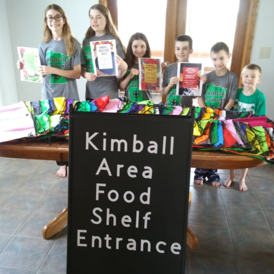 Six youth members standing behind a table of colorful assortment of bags and an a frame sign that reads, "Kimball Area Food Shelf Entrance."