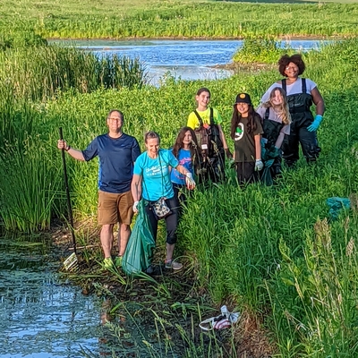 An adult with a group of youth standing next to a pond. The youth are wearing waders and work gloves.