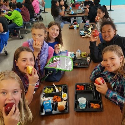 A group of seven elementary students smile while they eat apples and their school lunch in the cafeteria. 