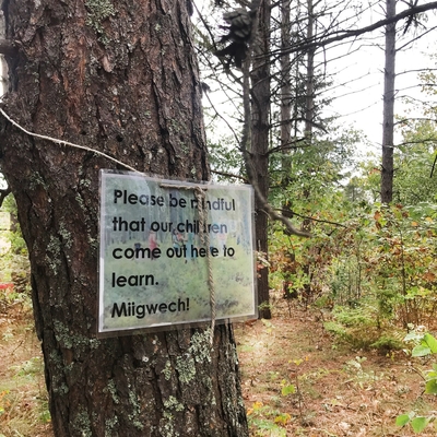 A tree with a sign hung that says "Please be mindful that our children come out here to learn. Miigwech!"