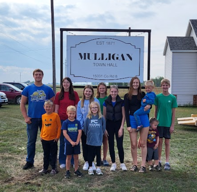 A group of youth of varying ages standing outside in front of a white sign that says, "Est. 1871, Mulligan Town Hall, 15031 Co. Rd. 8"