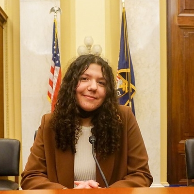 Teen sits at a table in the U.S. Capitol with two flags, a bookcase, and a door behind her.