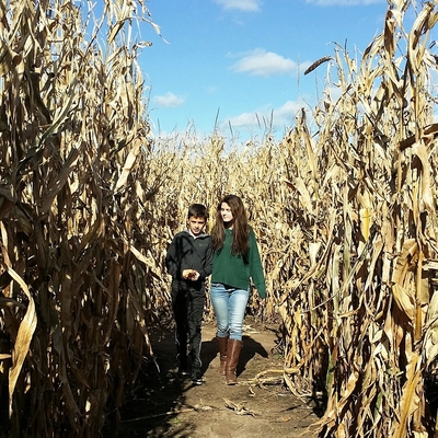 A tween boy and teen girl walk between rows of a corn maze under a blue sky.