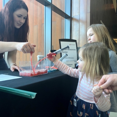 Woman leans over a table squeezing a tube in an aquarian filled with red dye while a small girl pumps from the other end to see how groundwater goes in and out of earth. Her big sister looks on.