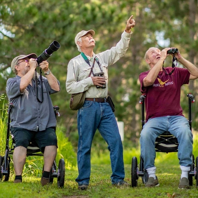 Three men out birding; two are seated on walker seats, one looks through a camera, one looks through binoculars, one points