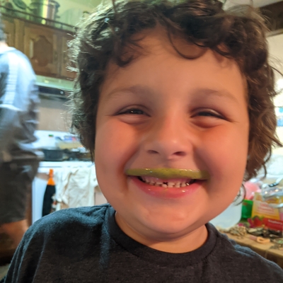 Smiling curly-haired boy shows off his green mustache. He is in a kitchen.