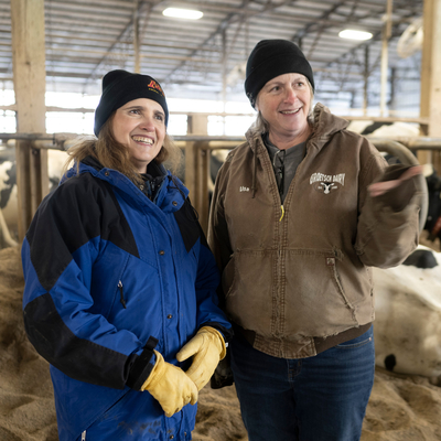 Dairy scientist and dairy farmer stand side by side in front of Holstein cows, smiling and gesturing