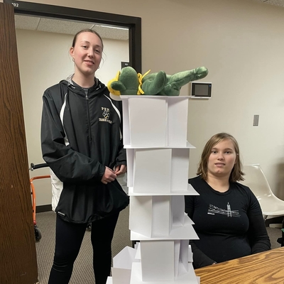 Two girls next to a notecard tower with a stuffed animal bear lying on top of the tower.