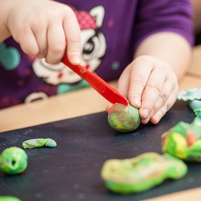 A close up have a child's hands cutting into green play doh with a red plastic knife. 