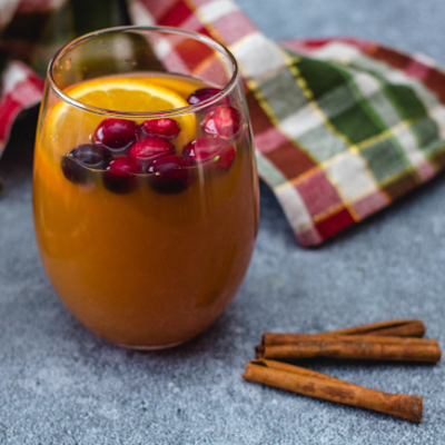 A cup of cider sitting on a gray counter with two cinnamon sticks next to it. In the cider is an orange slice and some berries.