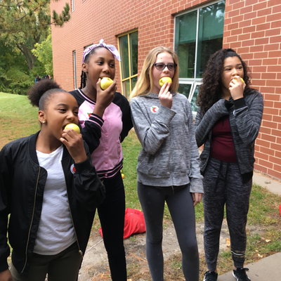A group of four students eat an apple outside of their school building. 