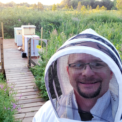 A man in a beesuit posing beside several beehives in a field.