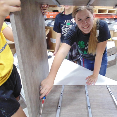 Victoria Shafer is working on some sort of woodworking project and smiling at the camera from behind a box that she is working on using a screwdriver