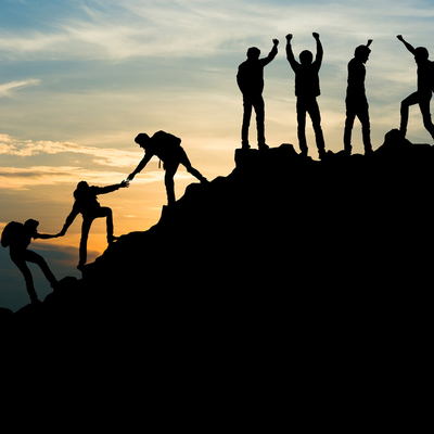 A silhouette of a group of people leading and helping each other climb a mountain as the sun sets.