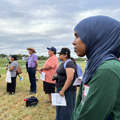 A line of people standing in a farm field watching a speaker.
