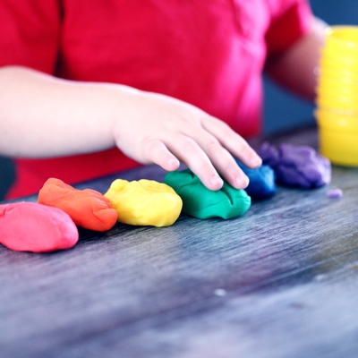 Playdoh balls in rainbow colors in a line on a table with a child's hand on one of the playdoh balls.