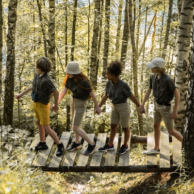 A line of youth holding hands while crossing a bridge in a wooded area.